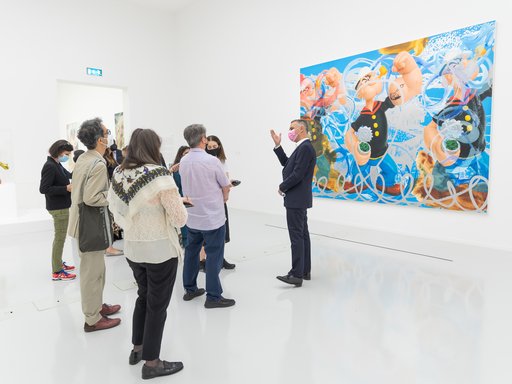 Massimiliano Gioni talking to a group of visitors in a gallery, standing in front of Jeff Koons' painting Triple Popeye