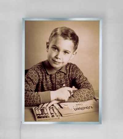 A black-and-white photograph of Jeff Koons as a child sitting at a table with a box of crayons and holding a large crayon in his right hand