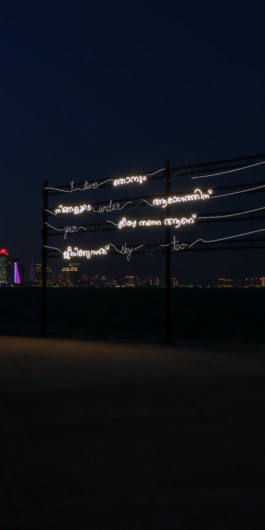 A neon art installation displaying multilingual text, against a backdrop of a dark cityscape on the waterfront at night.