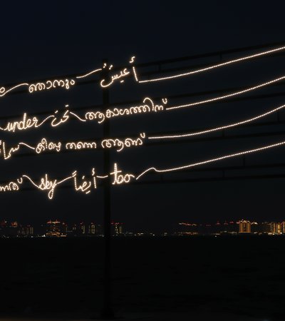 A neon art installation displaying multilingual text, against a backdrop of a dark cityscape on the waterfront at night.
