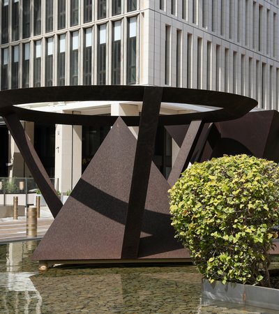 Two large abstract geometric sculptures in a shallow pool, with a modern building facade and greenery in the background.