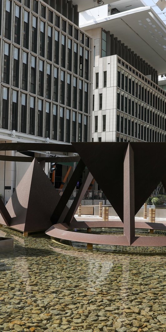 Two large abstract geometric sculptures in a shallow pool, with a modern building facade and greenery in the background.