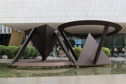 Two large abstract geometric sculptures in a shallow pool, with a modern building facade and greenery in the background.