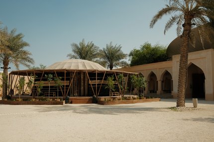 هيكل يشبه الخيمة مع أعمدة من الخيزران تحيط به في منتصف فناء محاط بأشجار النخيل.