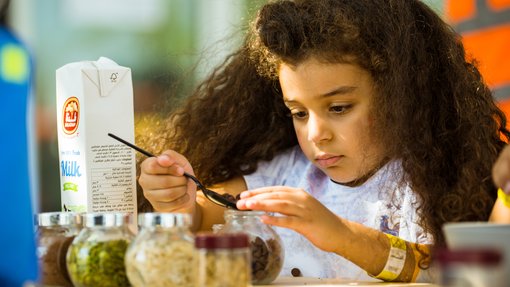 Little girl holding a spoonful of chocolate chips, next to her are various ingredients for baking