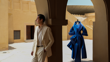 Benchellal in a cream-colored suit stands under an archway with a blue dress carved on a mannequin in a sunlit courtyard.
