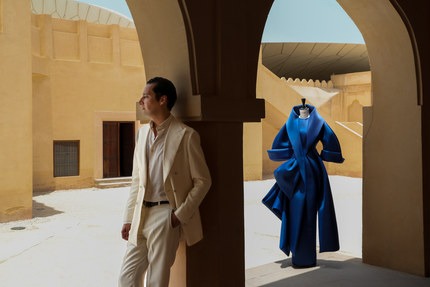 رجل يرتدي بدلة قشدية اللون يقف تحت ممر مقنطر مع فستان أزرق منحوت على عارضة أزياء في فناء مضاء بنور الشمس.