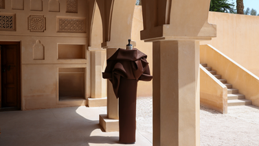 A brown sculpted dress on a mannequin standing in a sunlit courtyard with arched columns and ornate walls.