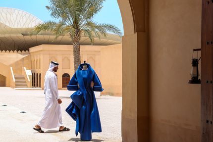 رجل يرتدي زيًا تقليديًا يمشي بجوار ثوب أزرق معروض على عارضة أزياء في فناء مضاء بنور الشمس مع أشجار النخيل.