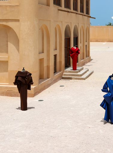 Benchellal in a white suit stands near four mannequins wearing colorful sculptural clothing outside a beige building on a sunny day.