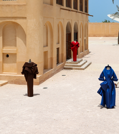 Benchellal in a white suit stands near four mannequins wearing colorful sculptural clothing outside a beige building on a sunny day.