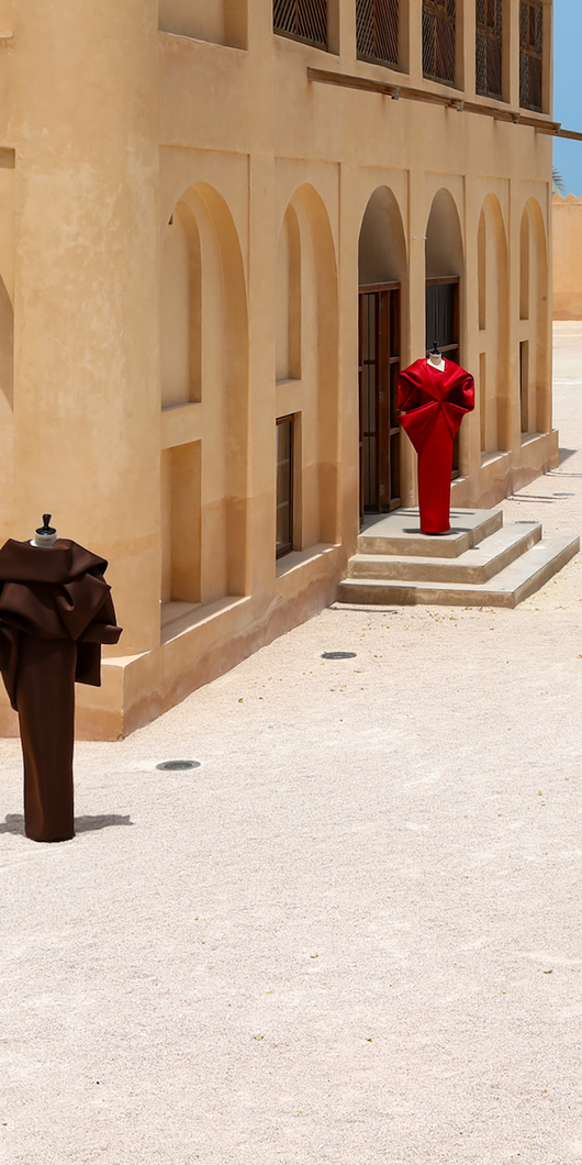 Benchellal in a white suit stands near four mannequins wearing colorful sculptural clothing outside a beige building on a sunny day.