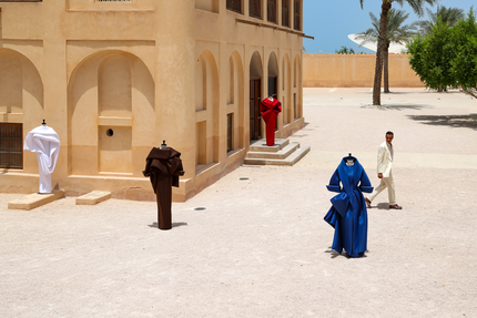 رجل يرتدي بدلة بيضاء يقف بالقرب من أربعة تماثيل عرض أزياء ترتدي ملابس منحوتة ملونة خارج مبنى بيج اللون في يوم مشمس.