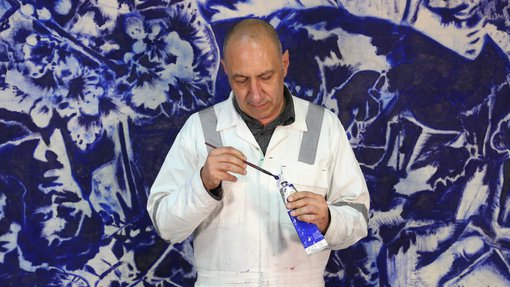 Visiting Artist Nedim Kufi standing in front of his painting, in his studio at the Fire Station, with oil paint and paint brush in hand.