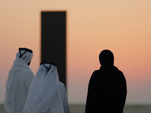 Three people in a desert landscape looking off into the sunset and two large black monoliths in the background.