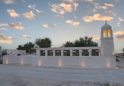 A white minaret and dome within a courtyard. The minaret is lit from the inside, against a blue sky with white clouds.