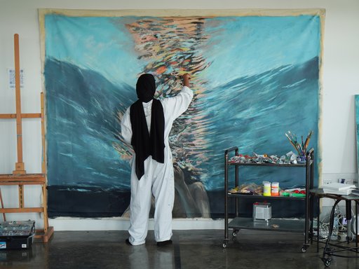 A girl with a black head cover and white overalls paints on a colourful canvas hung against a white wall.