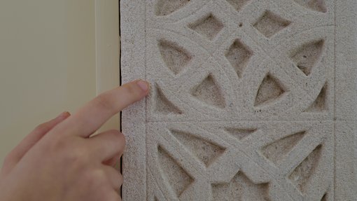 تلمس يد نمطًا هندسيًا منحوتًا بشكل معقد على سطح حجري فاتح اللون، مع جدار عادي مرئي على اليسار.