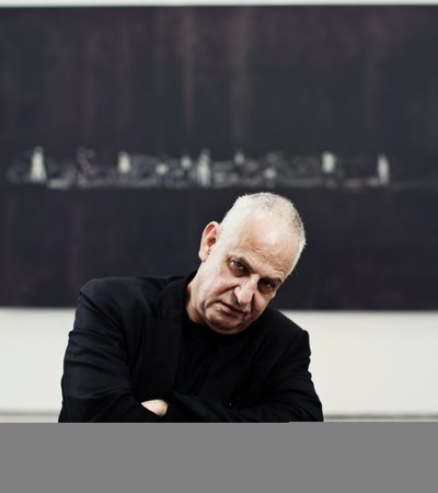 A portrait of Luc Tuymans framed by a black artwork in a museum while looking directly at the camera.