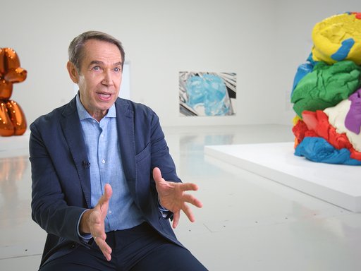 Jeff Koons at his exhibition, behind him are his public art displays of a orange balloon dog on the left and a play dough on the right