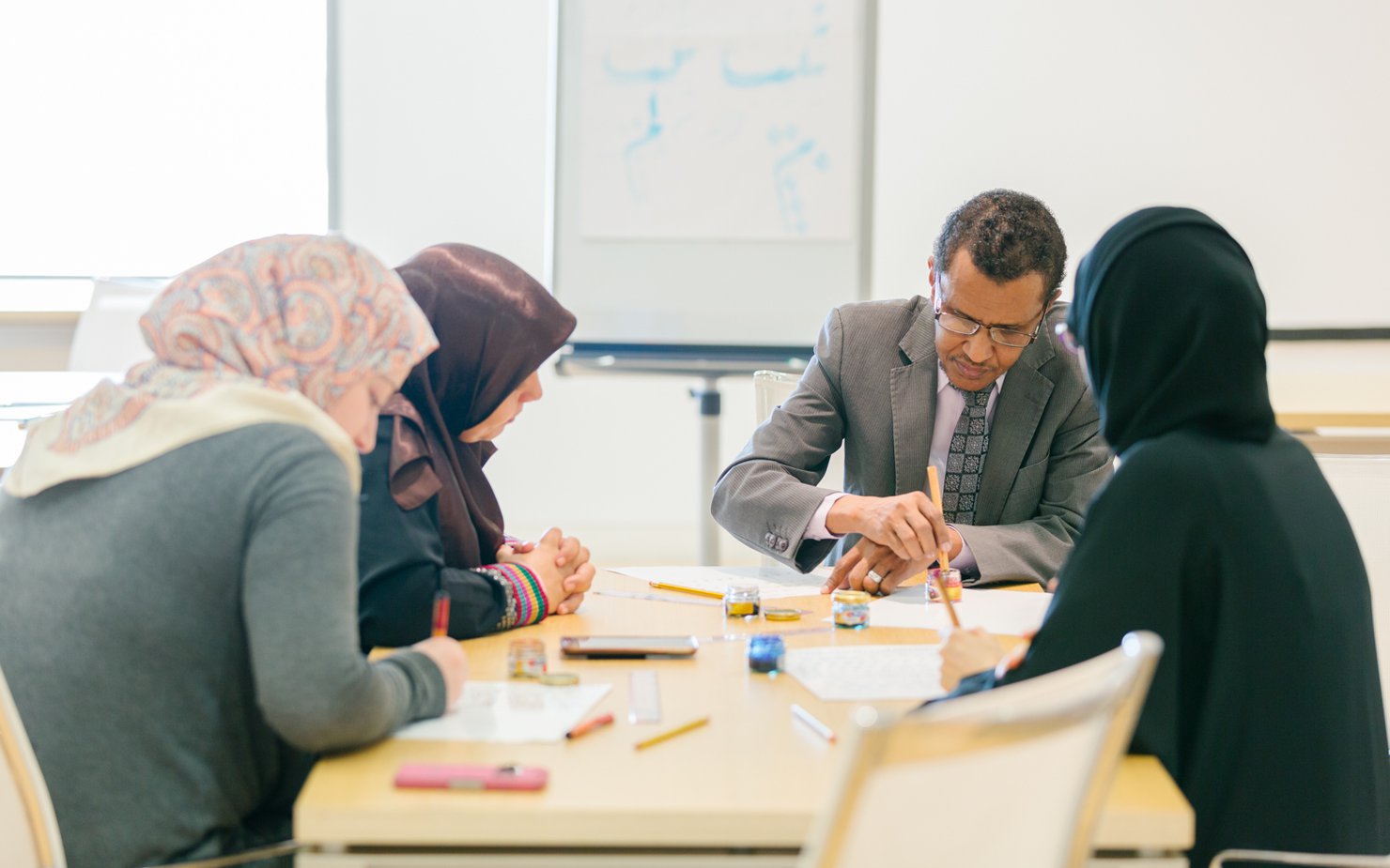 A man with a dip pen in his hand teaches calligraphy to three students circled around him over a desk