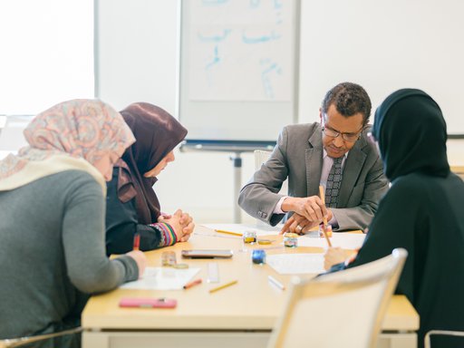 A man with a dip pen in his hand teaches calligraphy to three students circled around him over a desk